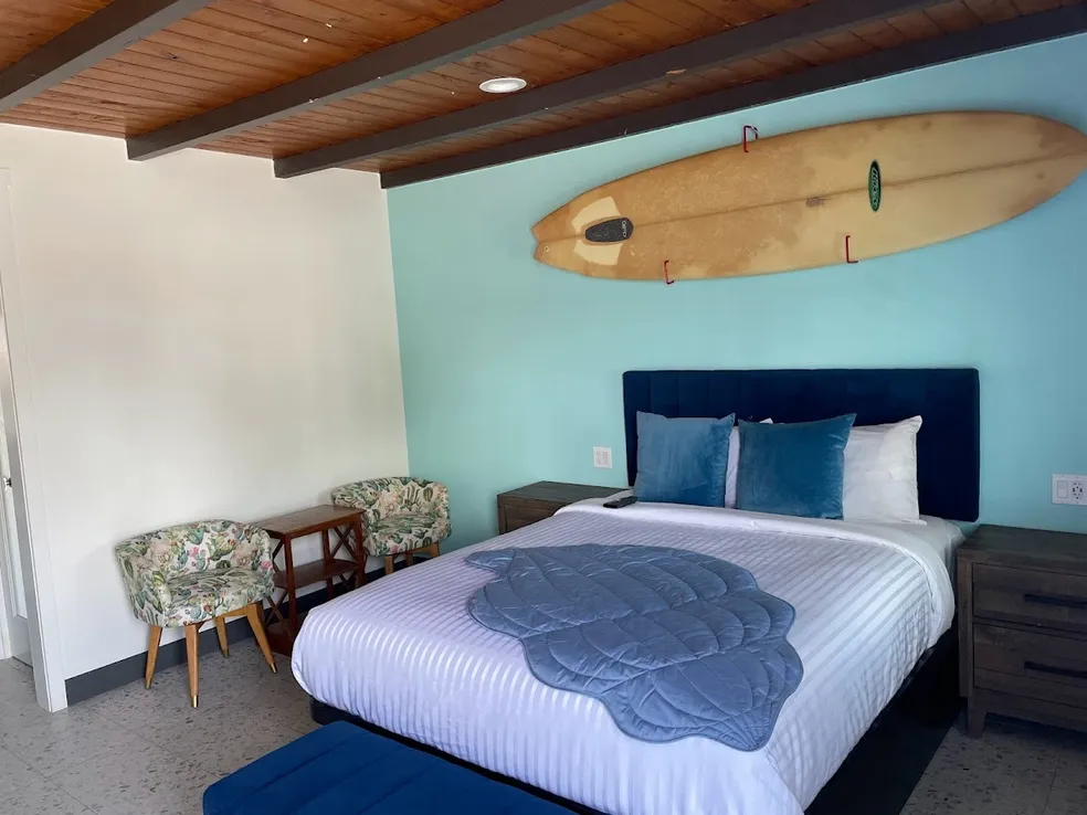 A Calafia Inn room with a vintage wooden surfboard mounted above the bed, a mint accent wall and a sitting area with two patterned chairs