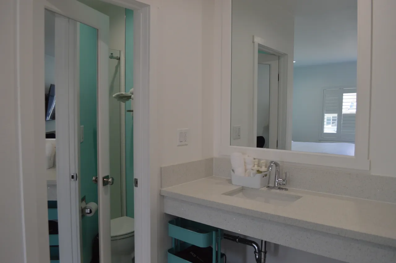 Deluxe room bathroom vanity with a white countertop and sink, an open door to a turquoise toilet room and a mirror reflecting the bed
