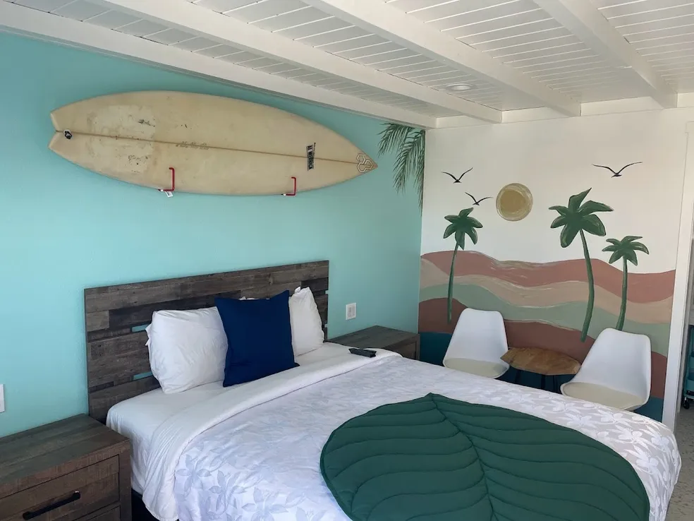 Standard room with a turquoise accent wall, a surfboard mounted above the bed and a hand-painted mural of dunes and palms