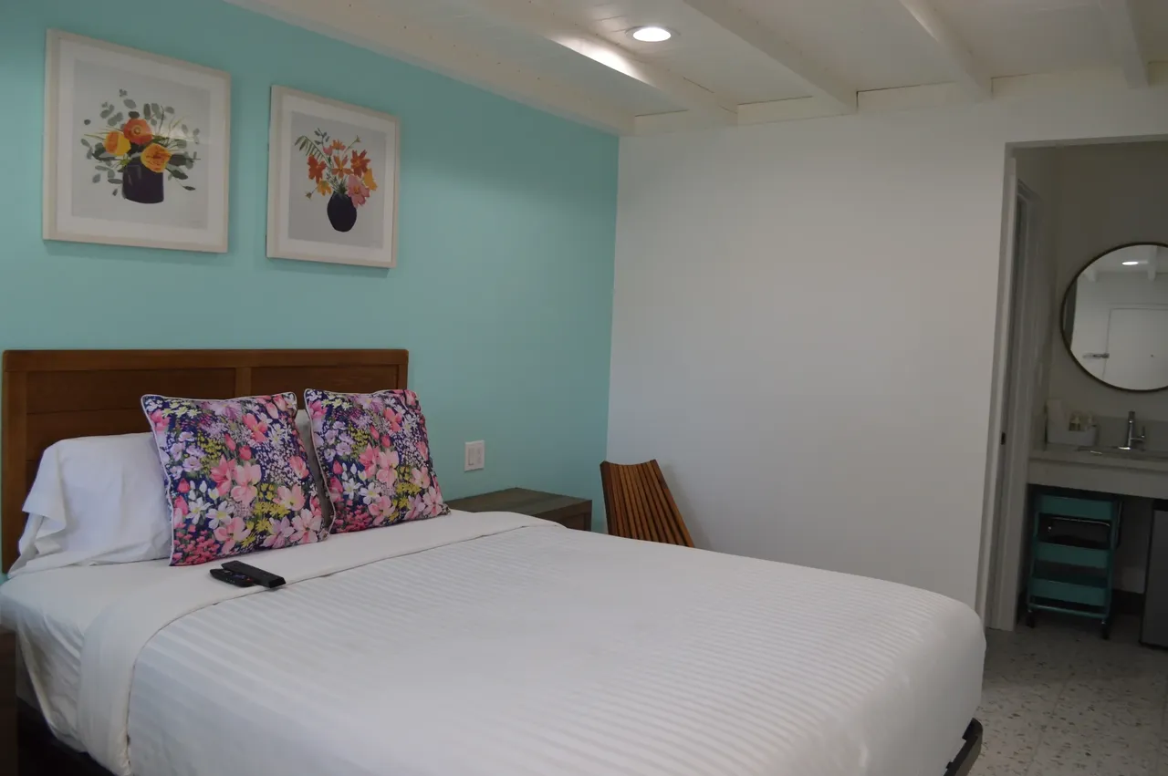 Standard room bed with floral pillows, a turquoise accent wall and a vanity visible through the doorway